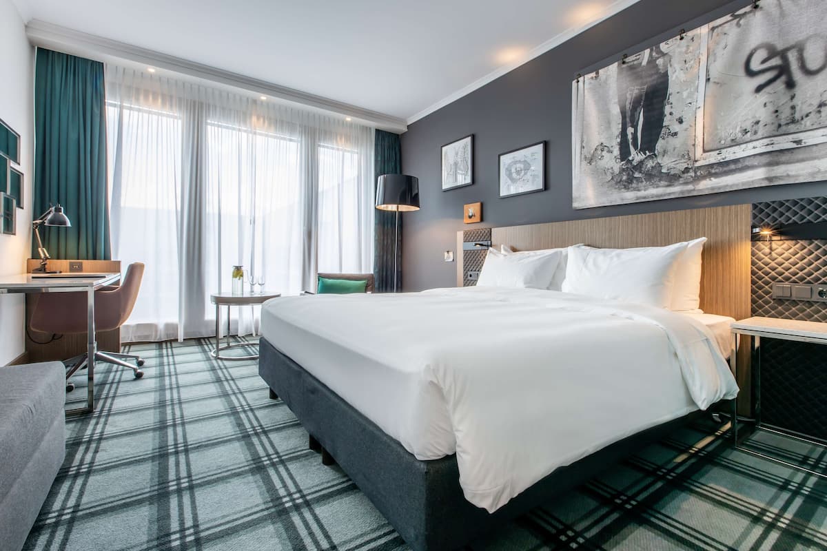 A hotel room with a bed, desk, chair, and large windows with curtains.