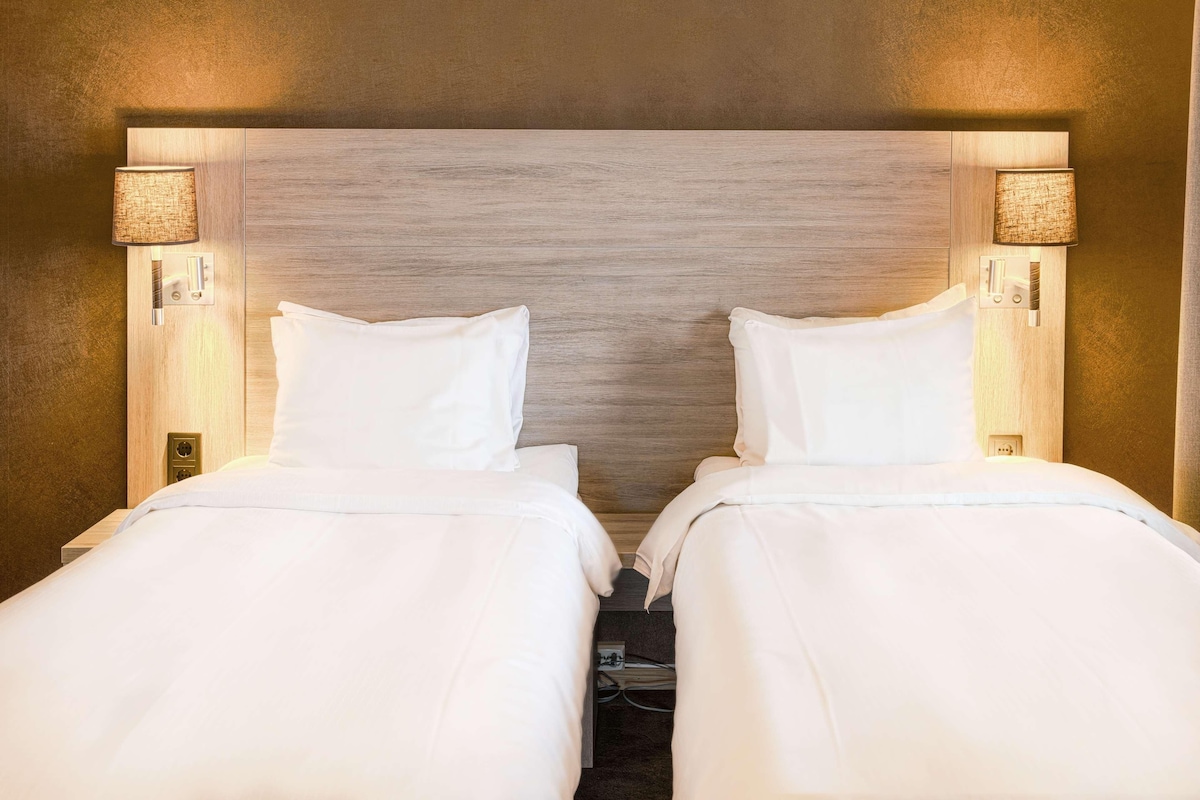 A hotel room with two single beds, each with a white bedspread and pillows, flanked by wall-mounted lamps.