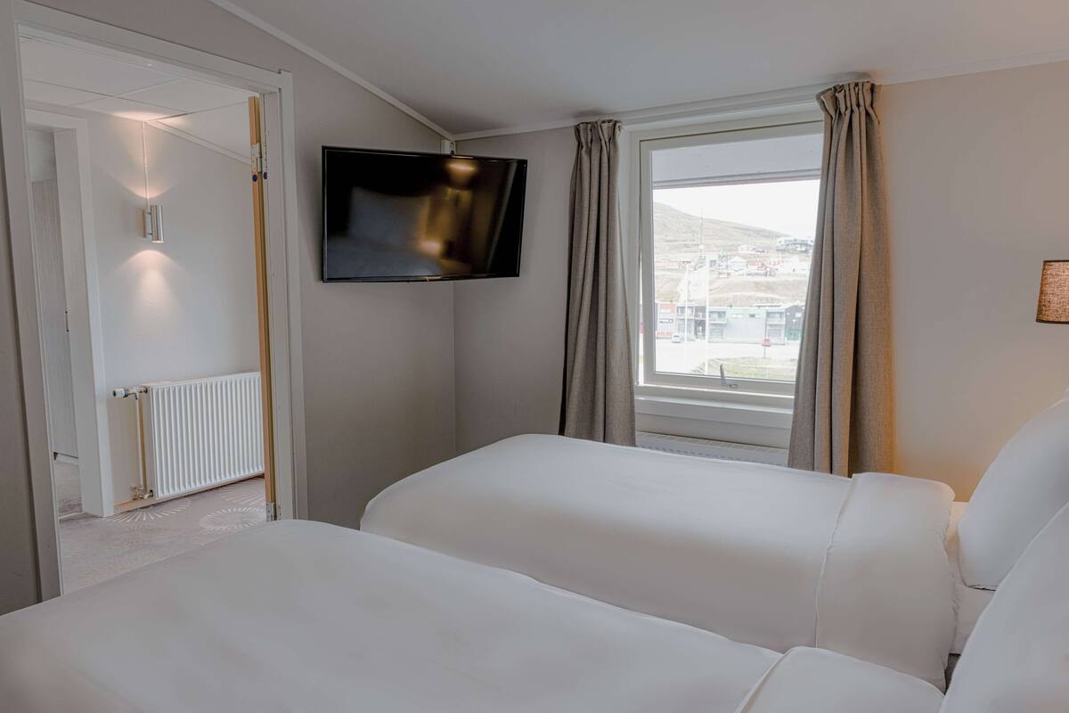 A hotel room with a bed, a TV mounted on the wall, a window with curtains, and a view of the outside.