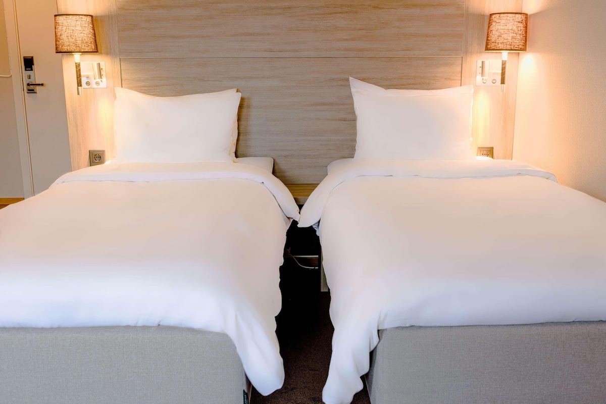 A hotel room with two beds, each with white bedding and a wooden headboard. There are wall-mounted lamps on either side of the beds.