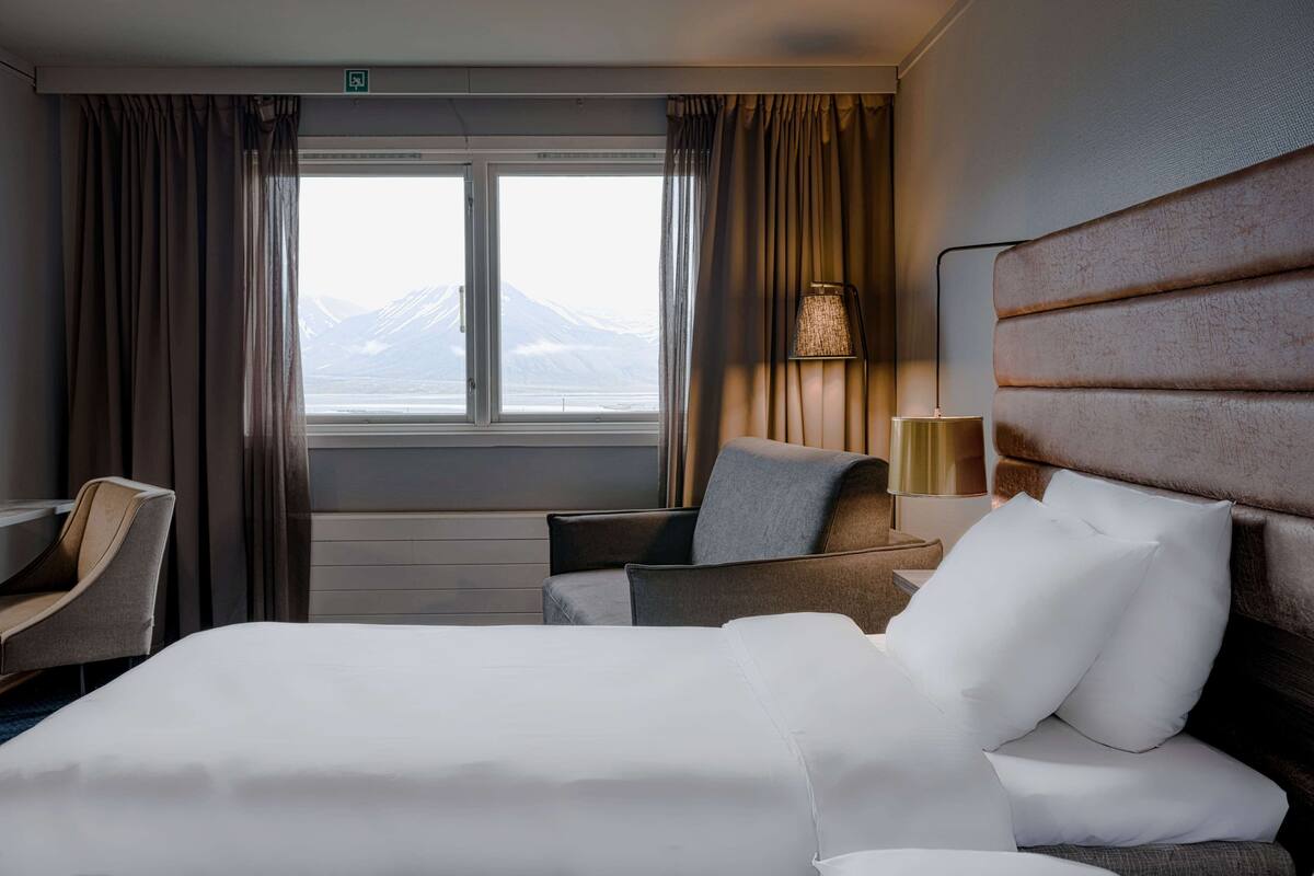 A hotel room with a bed, a desk, a chair, a lamp, and a view of a mountain through the window.