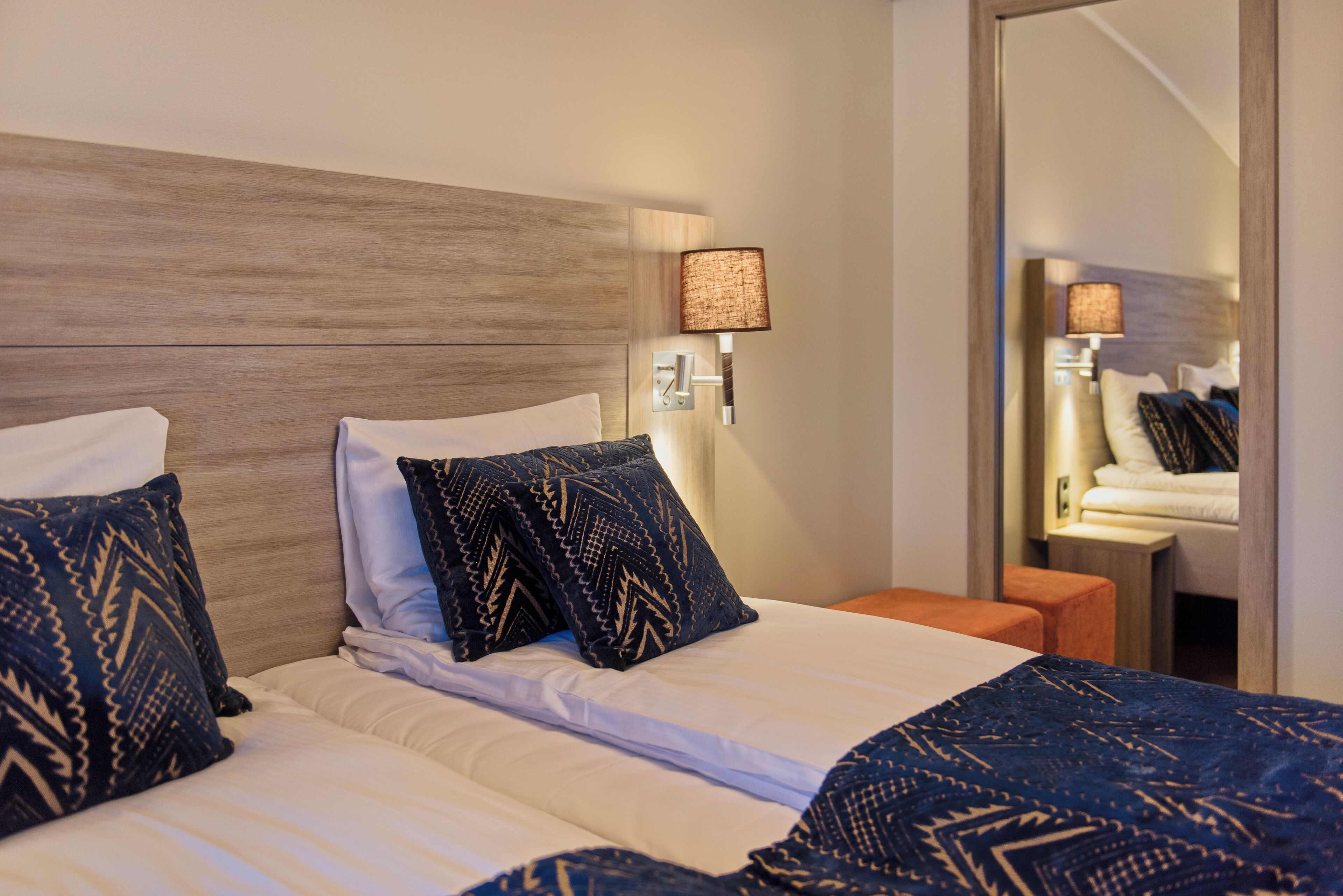 A hotel room with a wooden headboard, two beds with patterned pillows, a nightstand, a mirror, and a wall-mounted lamp.