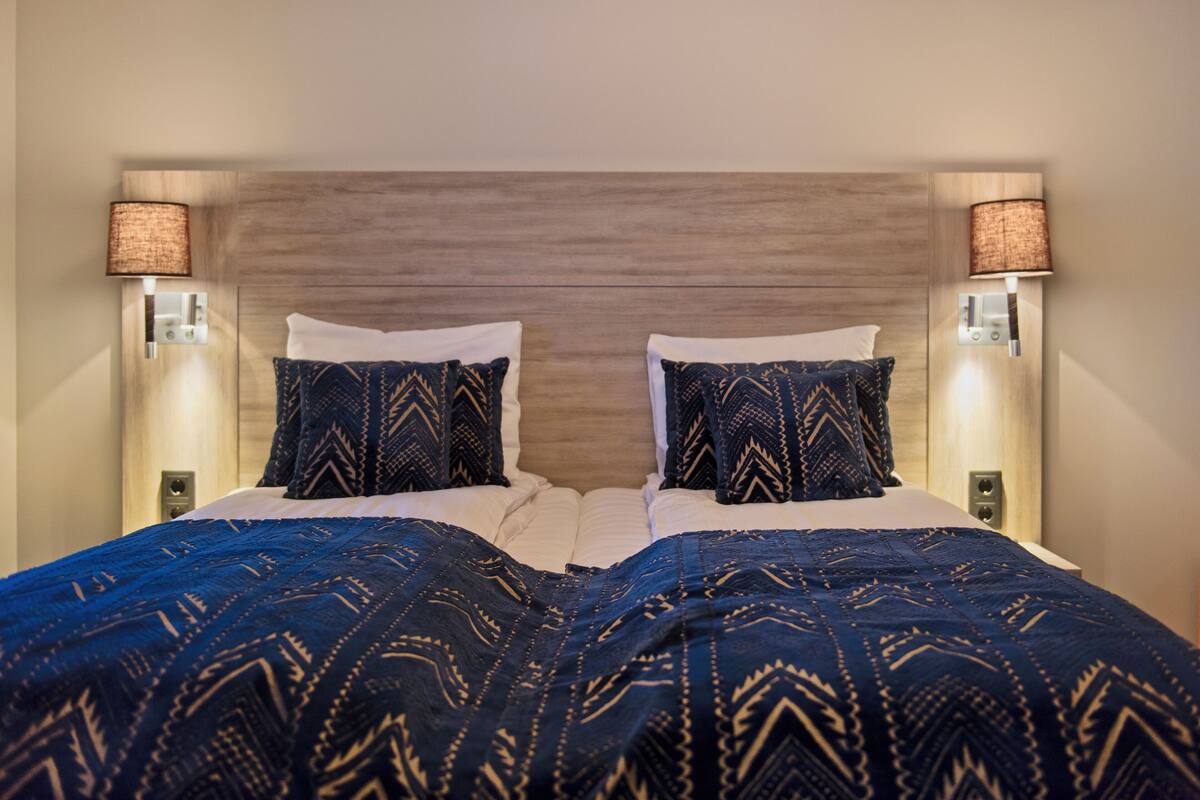 A hotel room with a wooden headboard, two bedside lamps, a patterned blue bedspread, and white pillows.