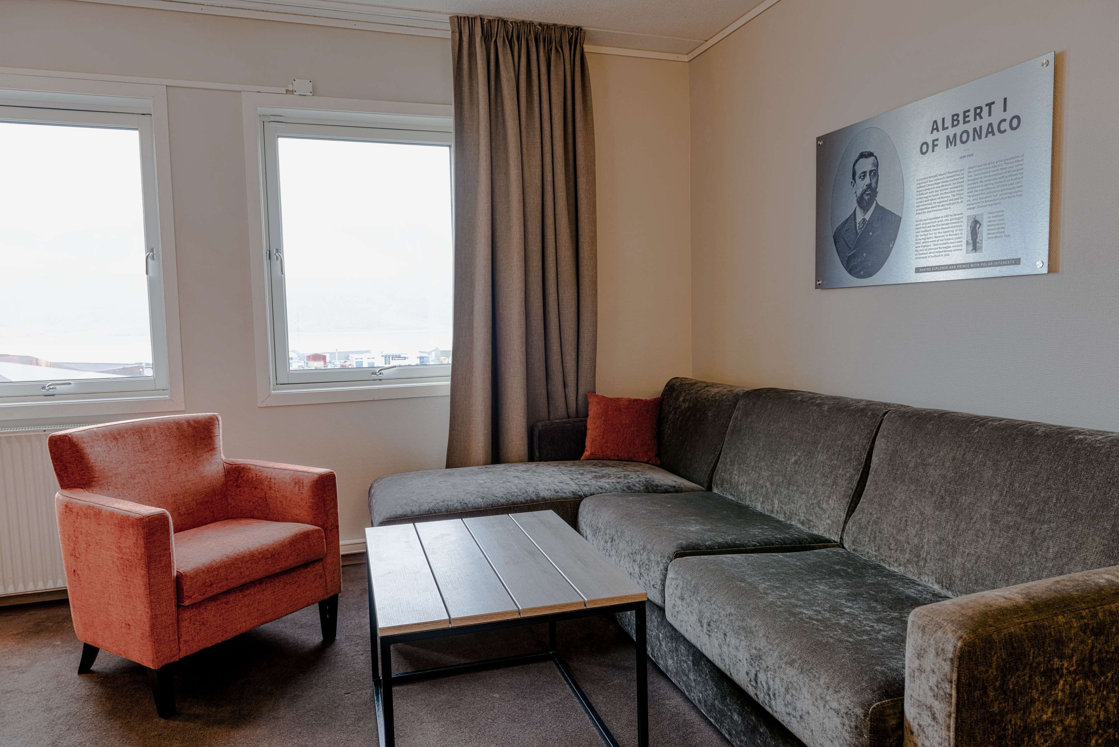 A living room with a grey sofa, a red armchair, and a wooden coffee table. There is a window with curtains, and a framed picture of Albert I of Monaco on the wall.