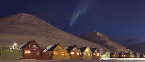 File di cabine luminose con tetti imbiancati dalla neve, sullo sfondo una montagna e un cielo notturno sereno.
