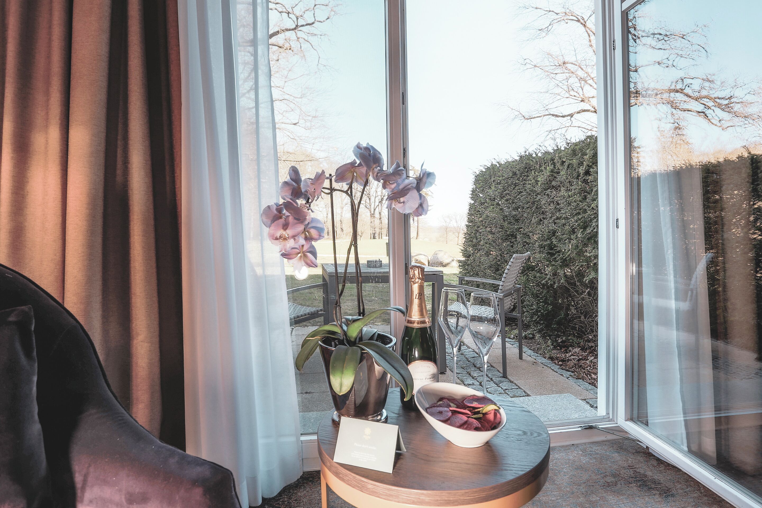 A room with a large window, a chair, a table with a bottle and glasses, and a bowl of fruit.