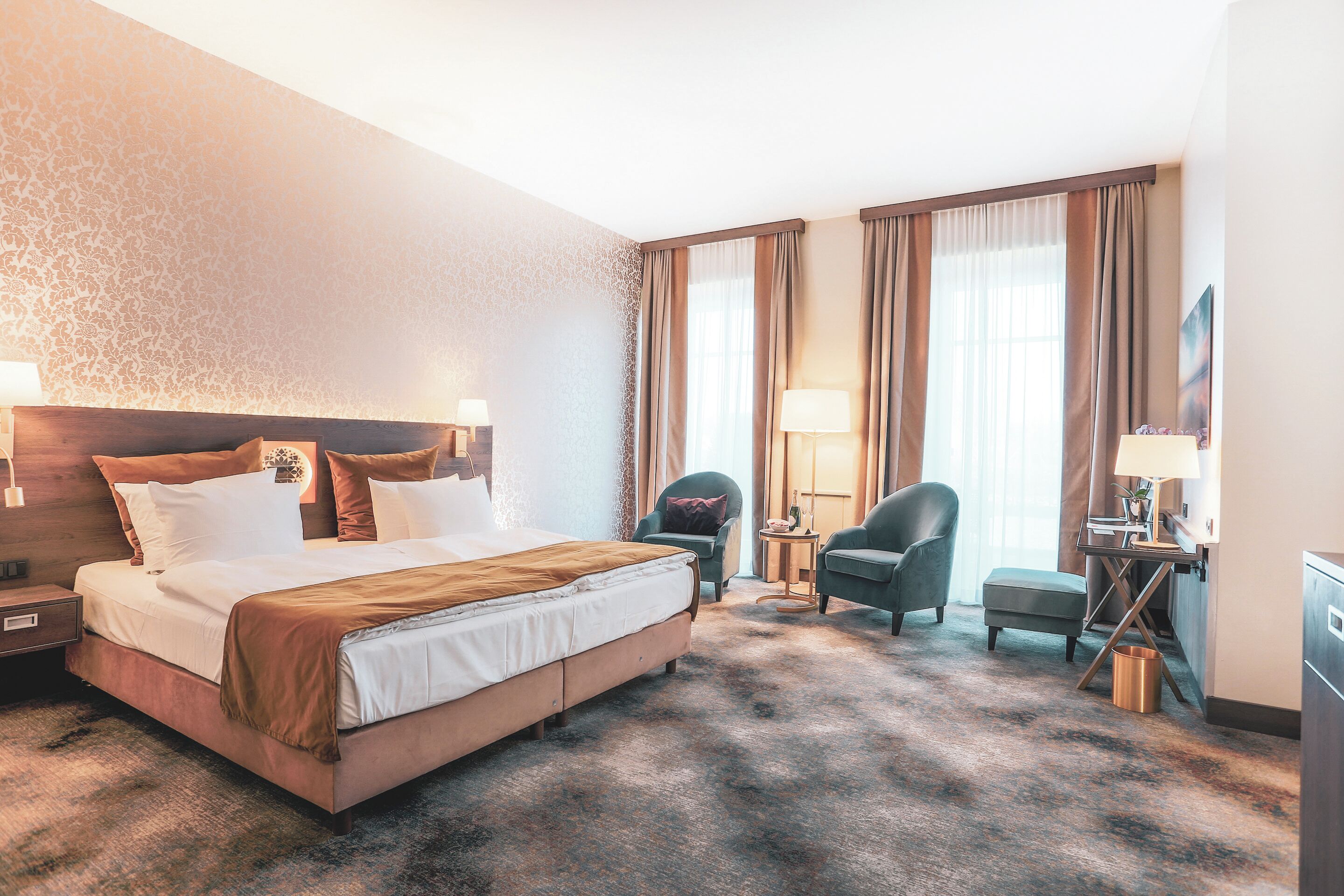 A hotel room with a large bed, two armchairs, a desk with a lamp, and a large window with curtains.