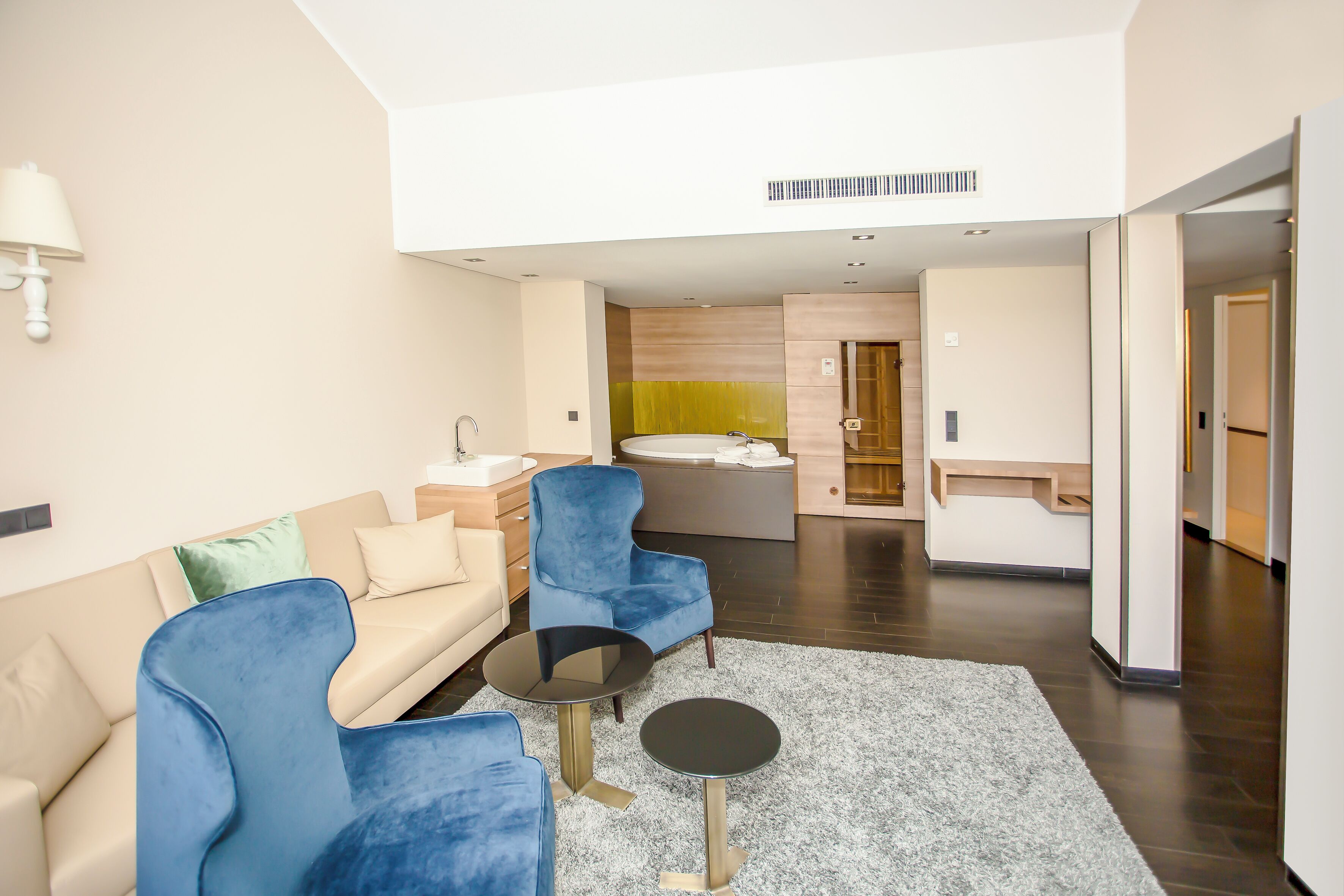 Une chambre d’hôtel moderne comprenant un canapé, deux fauteuils, une petite table ronde, une salle de bain avec baignoire et un couloir menant à une autre chambre.