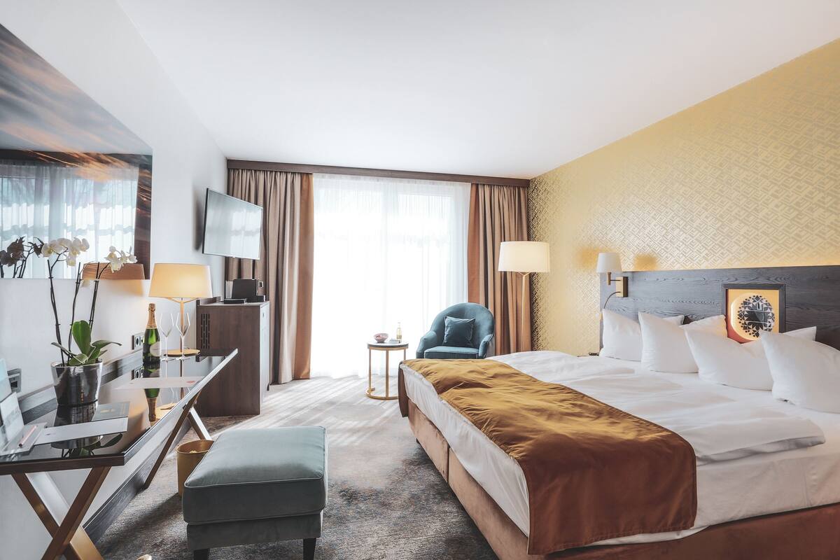 A hotel room with a bed, a desk, a chair, a TV, and a small table with a bottle and glasses.
