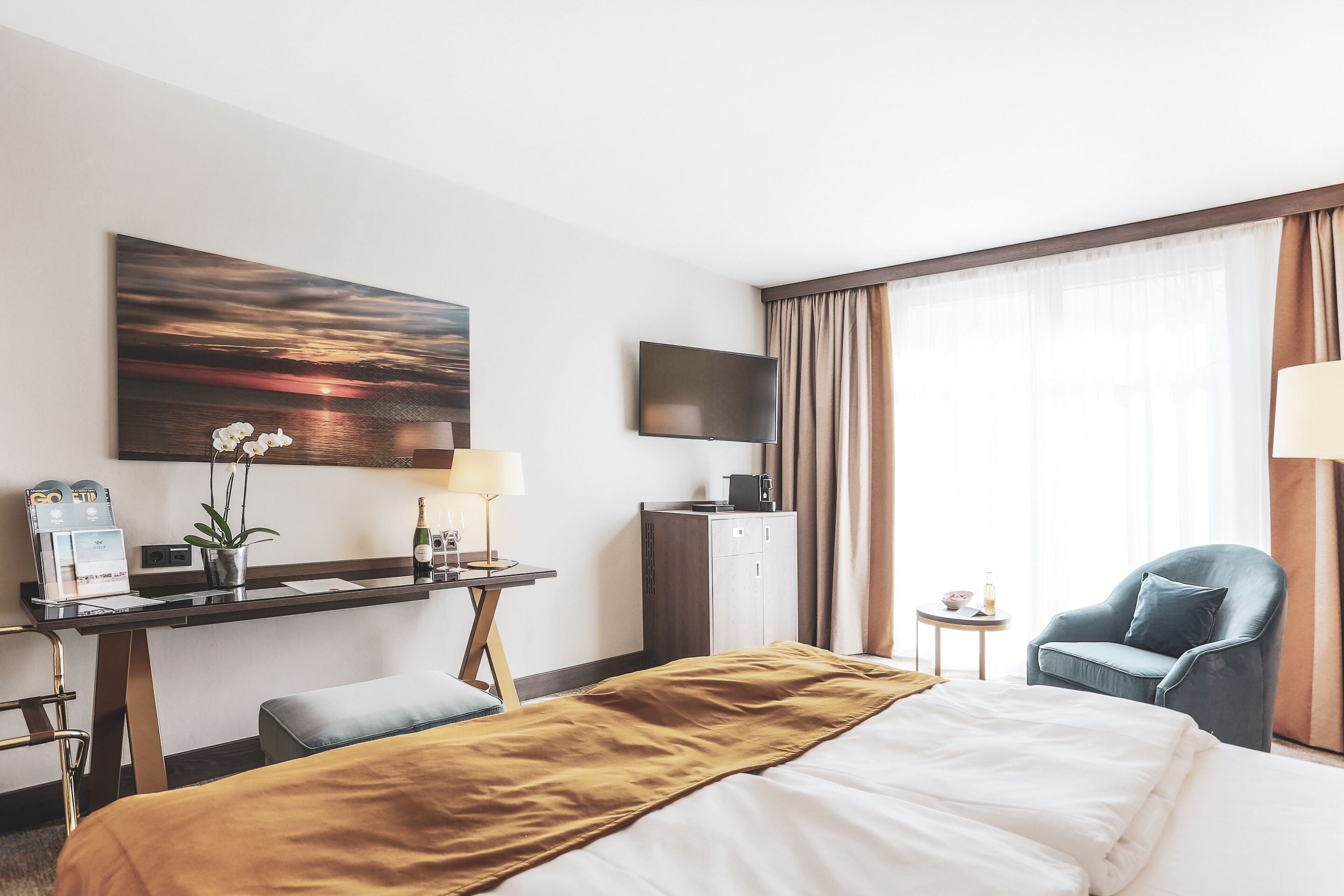 A hotel room with a bed, a desk with a lamp, a chair, a TV, and a painting on the wall.