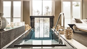 Een modern interieur met een centrale glazen trap, een zithoek met een bank en fauteuils, en decoratieve objecten op een console tafel.