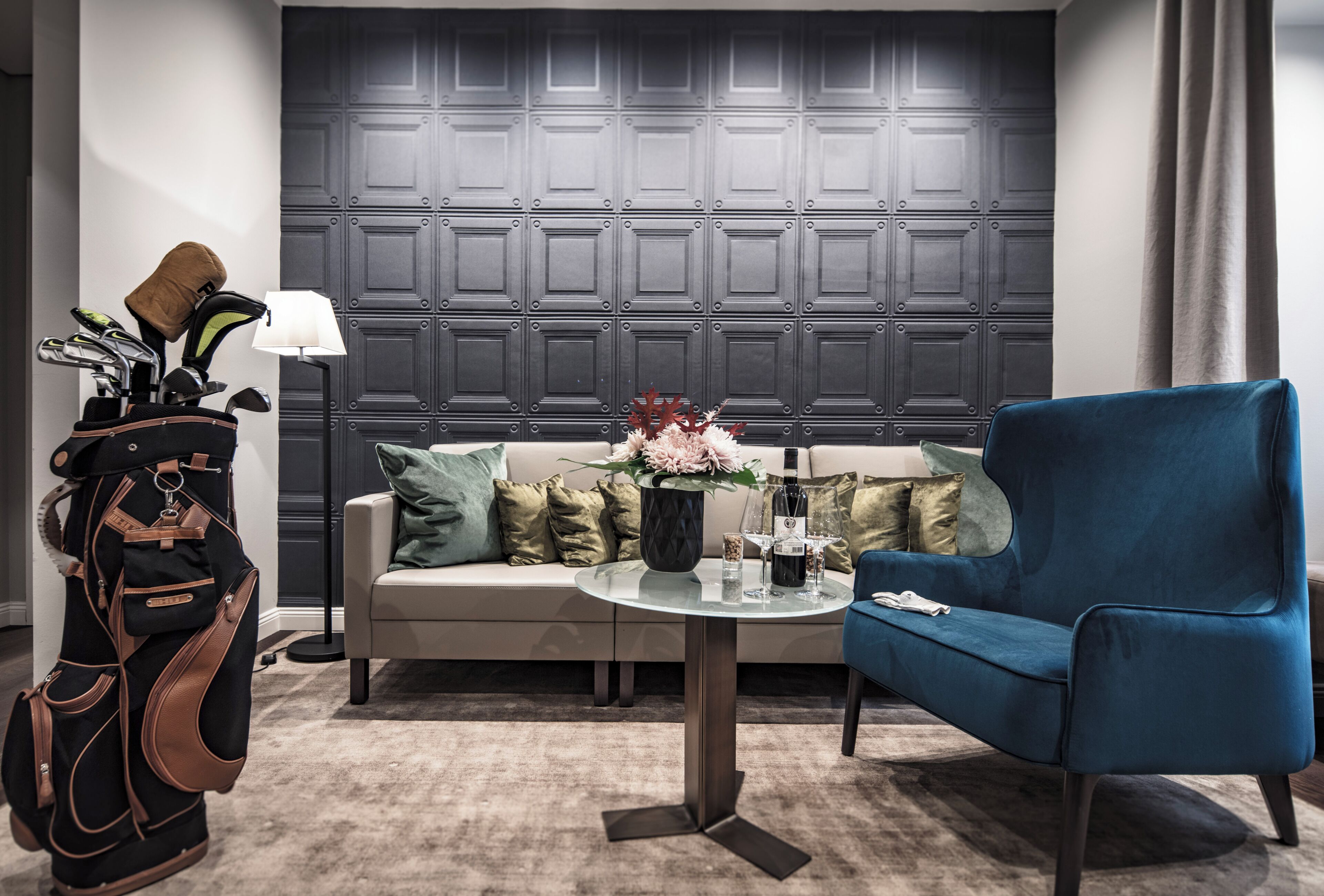 A modern living room with a sofa, a blue armchair, a coffee table with a bottle and glasses, and a golf bag.
