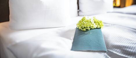 Una cama bien tendida con ropa de cama y almohadas blancas, una almohada azul con texto y un pequeño ramo de flores.