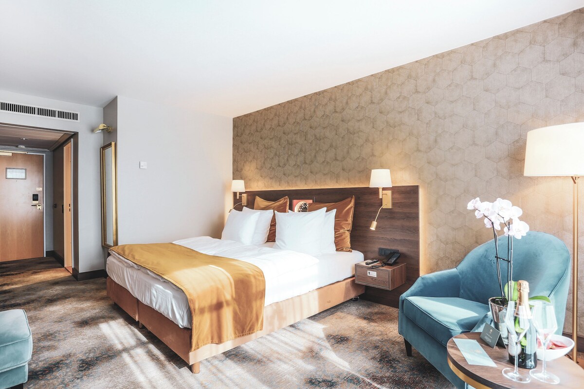 A hotel room with a bed, bedside table, chair, and a small table with a bottle and glasses.