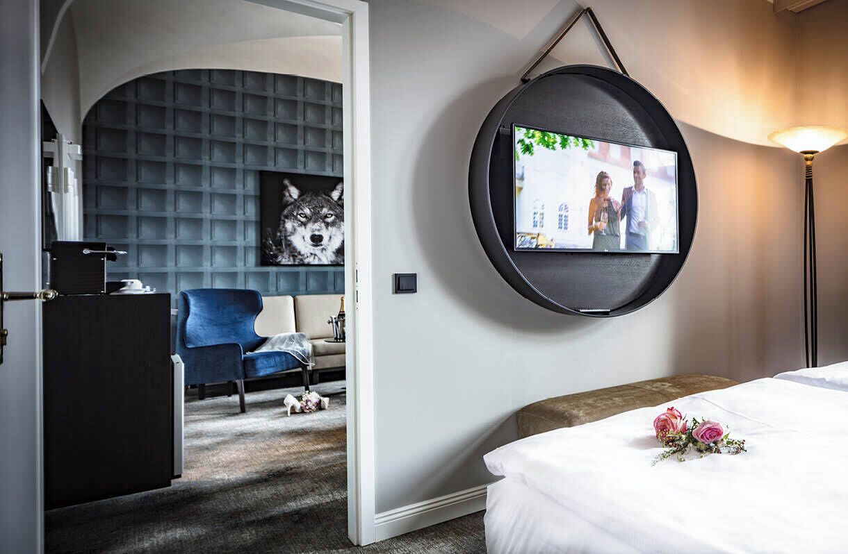 Quarto de hotel com uma cama, televisão na parede, uma cadeira azul, uma mesinha e um quadro decorativo de um lobo.