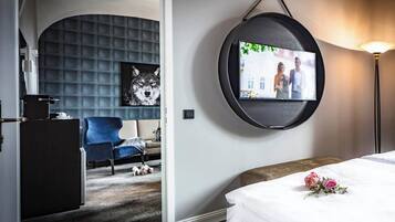 Quarto de hotel com uma cama, televisão na parede, uma cadeira azul, uma mesinha e um quadro decorativo de um lobo.