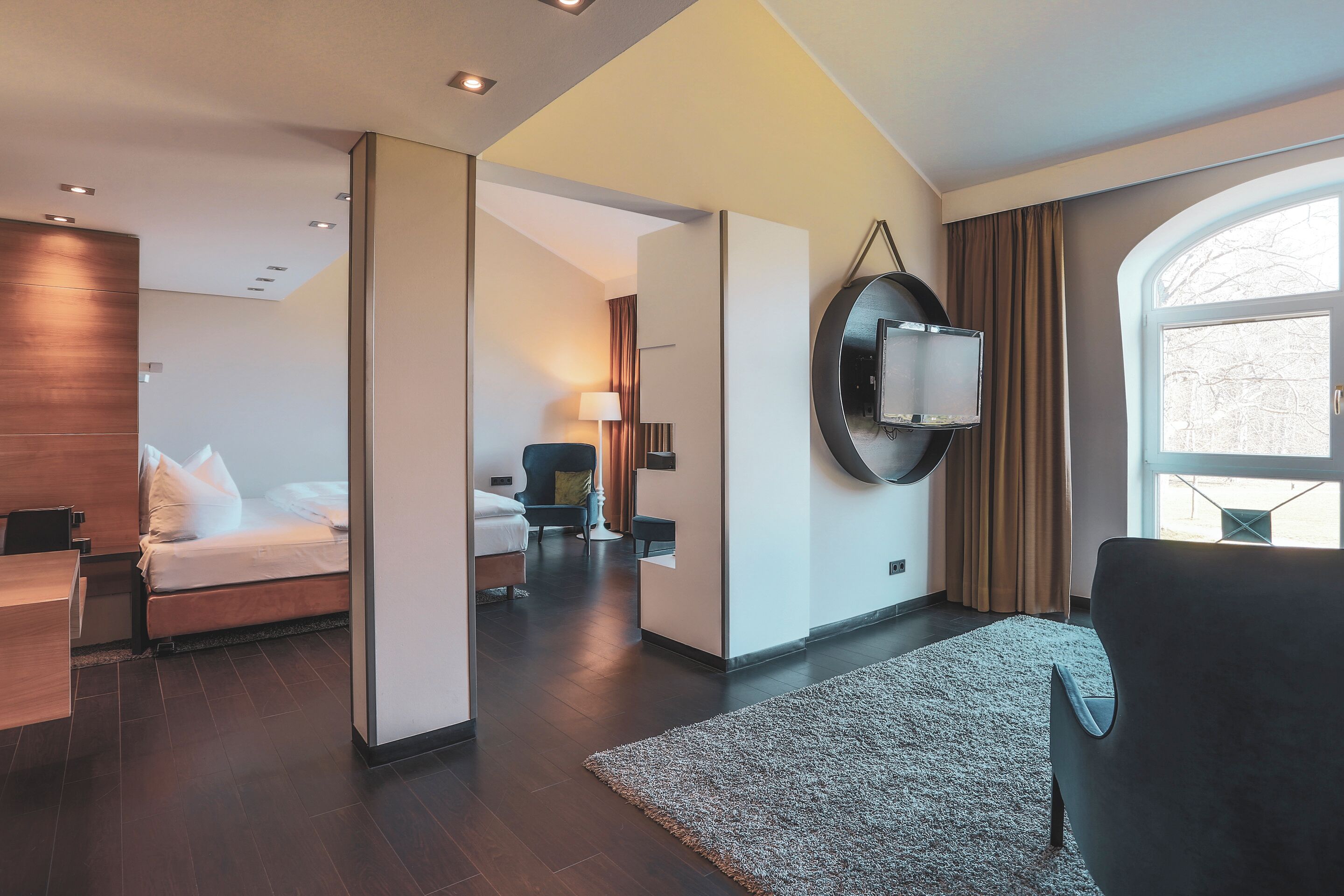 A modern hotel room with a bed, a desk, a chair, a large mirror, and a view of the outdoors.