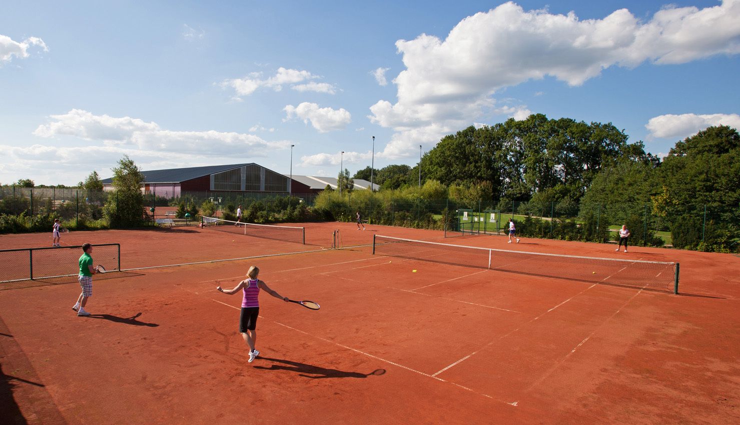 Een tennisbaan met spelers, een gebouw op de achtergrond en bomen eromheen.