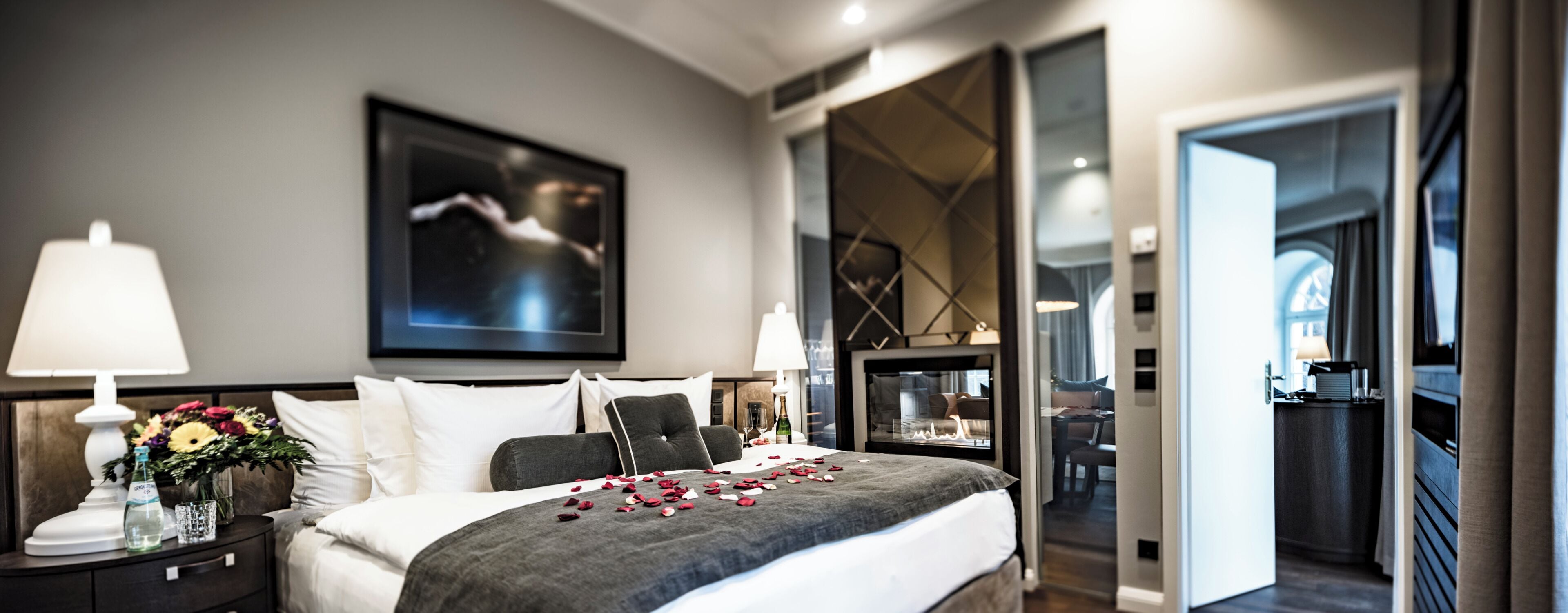 Una habitación de hotel moderna con una cama grande, una mesita de noche con lámpara, un jarrón de flores y un cuadro en la pared.