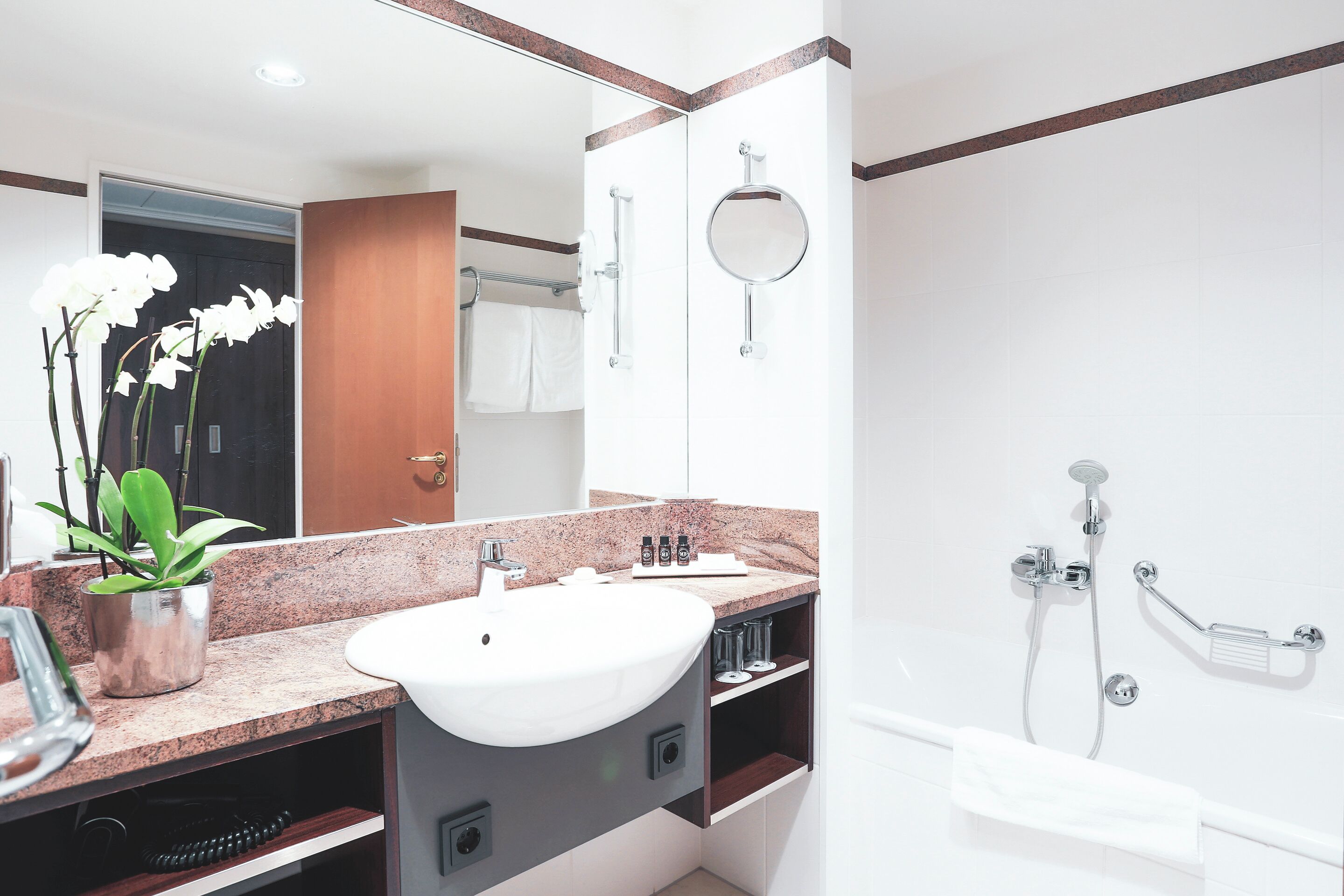 Une salle de bain moderne avec un lavabo blanc, un miroir rond, une douchette à main et une orchidée en pot.