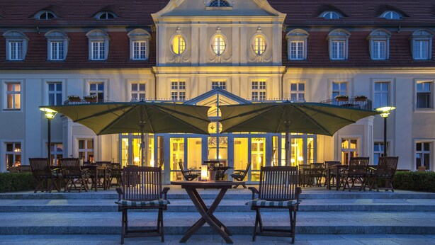 Een gebouw met een brede ingang, terraszitplaatsen met parasols en een goed verlicht interieur.