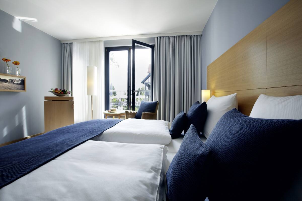 A hotel room with two beds, a view of the outdoors, a small table with a chair, and a window with curtains.