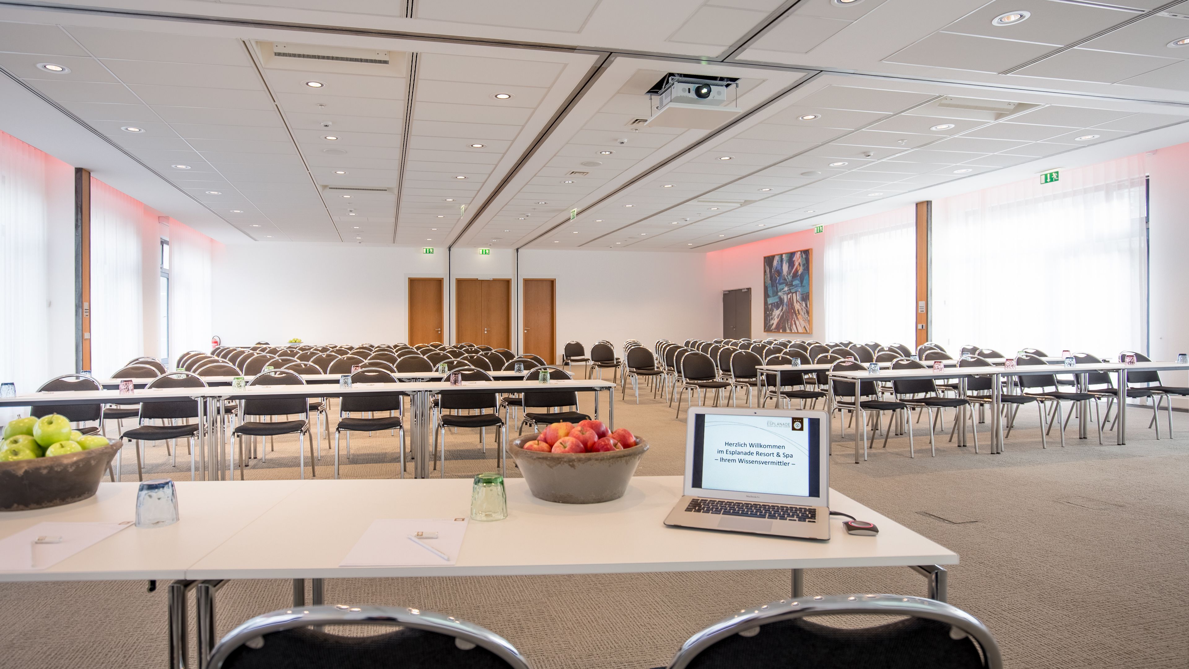 Un salón de conferencias con filas de mesas y sillas, una computadora portátil, un frutero y un mensaje de bienvenida en una pantalla.