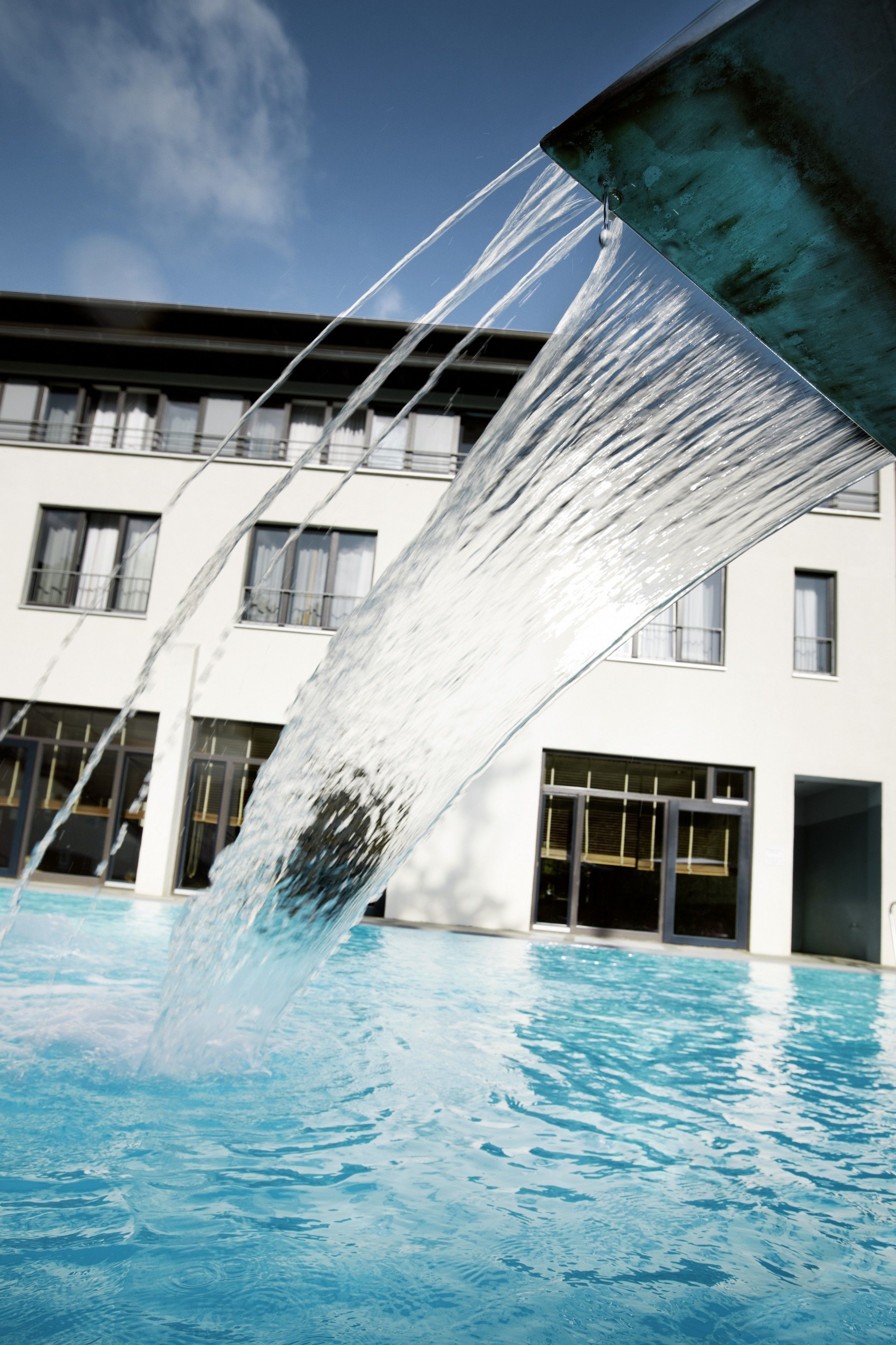 Uma piscina com água caindo de uma estrutura, rodeada por um edifício com várias janelas.