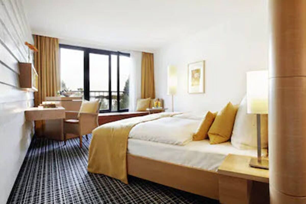 A hotel room with a bed, a desk, a chair, a lamp, and a large window.