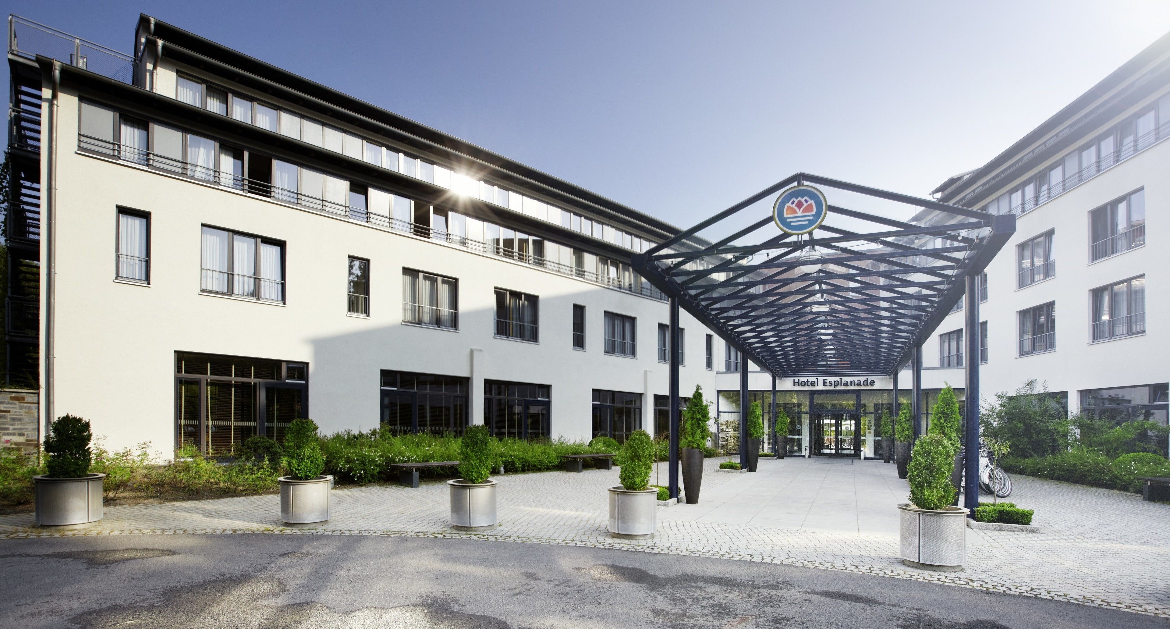 Une entrée d’hôtel moderne, dotée d’une allée couverte, de plantes en pot et surplombant un ciel dégagé.