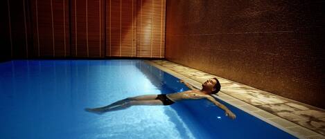 A person floating in a swimming pool with wooden blinds and a tiled wall in the background.