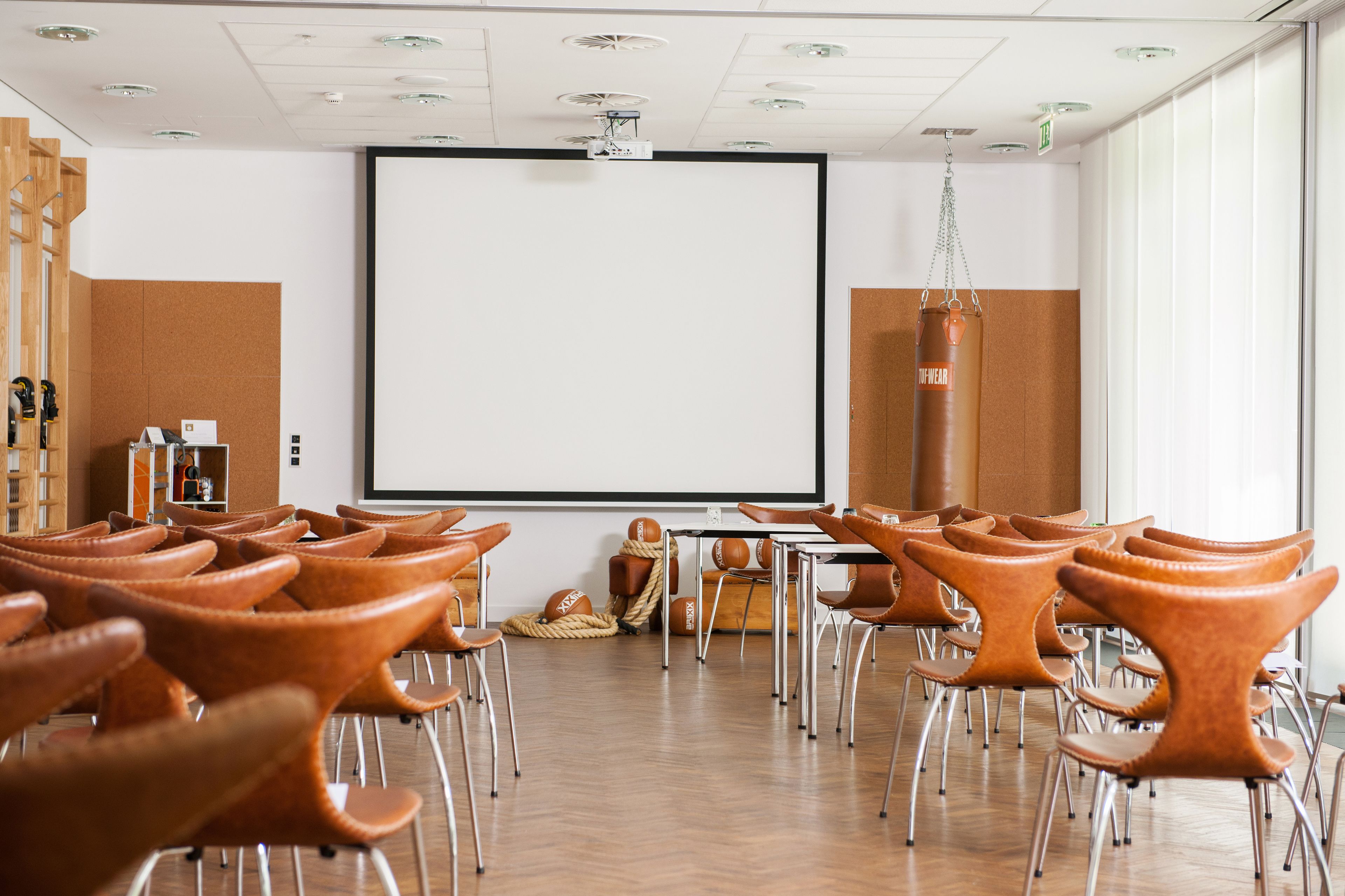 Un salón de conferencias con una pantalla grande, suelo de madera y sillas de cuero marrón.