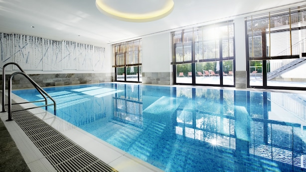 An indoor swimming pool with clear water, a metal ladder, and large windows with blinds.