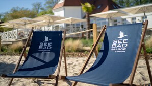 Two beach chairs with "SEE BAD SAAROW by Esplanade" printed on them, placed on sandy ground with umbrellas and a building in the background.
