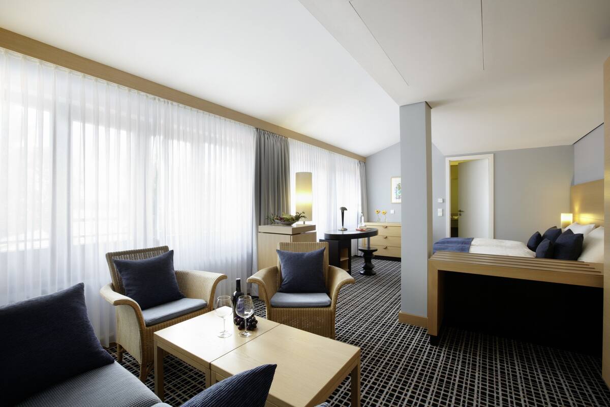 A hotel room with a bed, two armchairs, a small table, a minibar, and a view of the hallway.