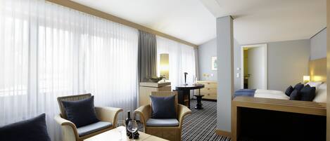 Habitación de hotel con una cama, dos sillones, una mesita, un minibar y vistas al pasillo.