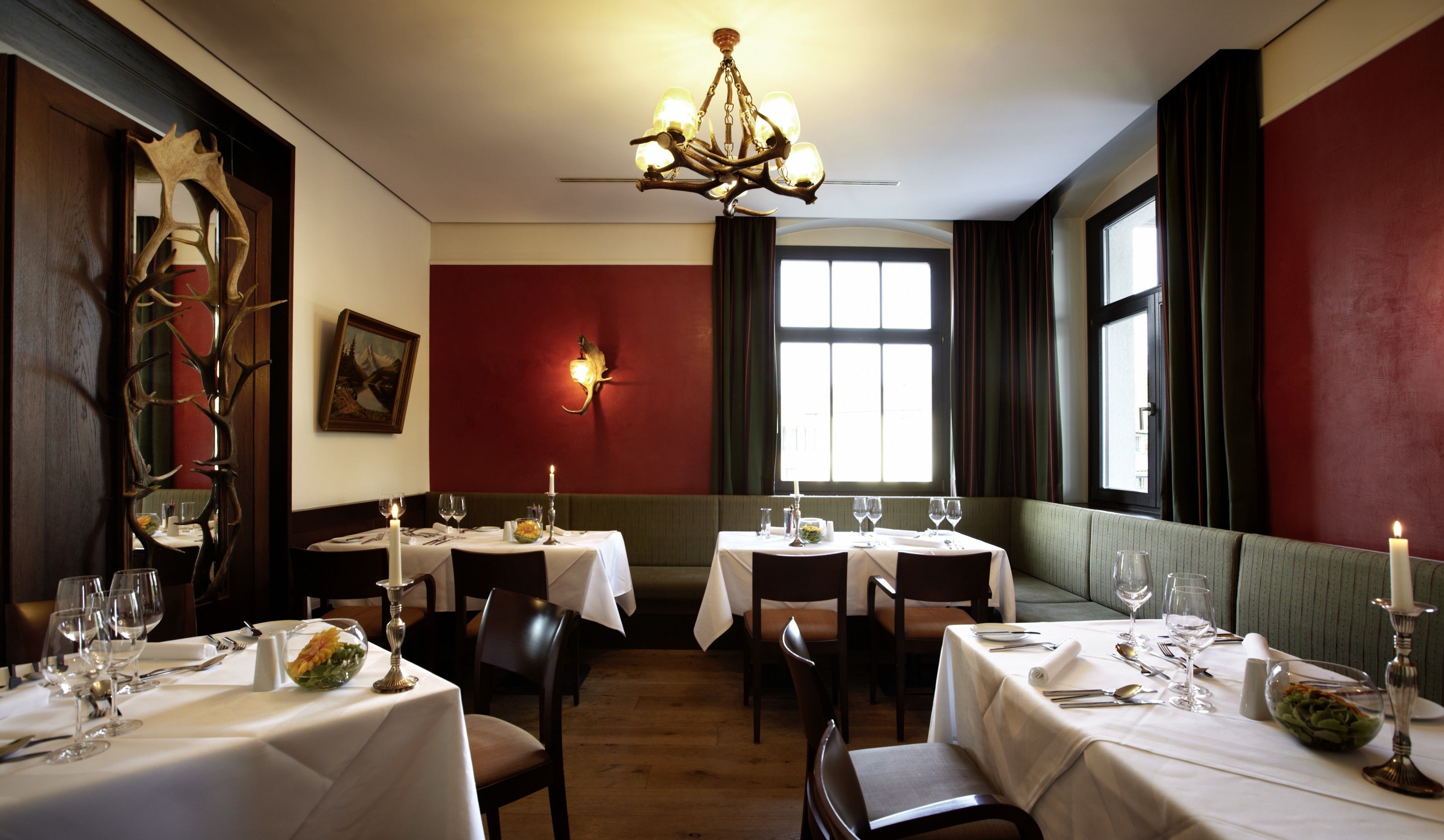 A dining area with tables set for four, a chandelier, a painting on the wall, and a window with curtains.
