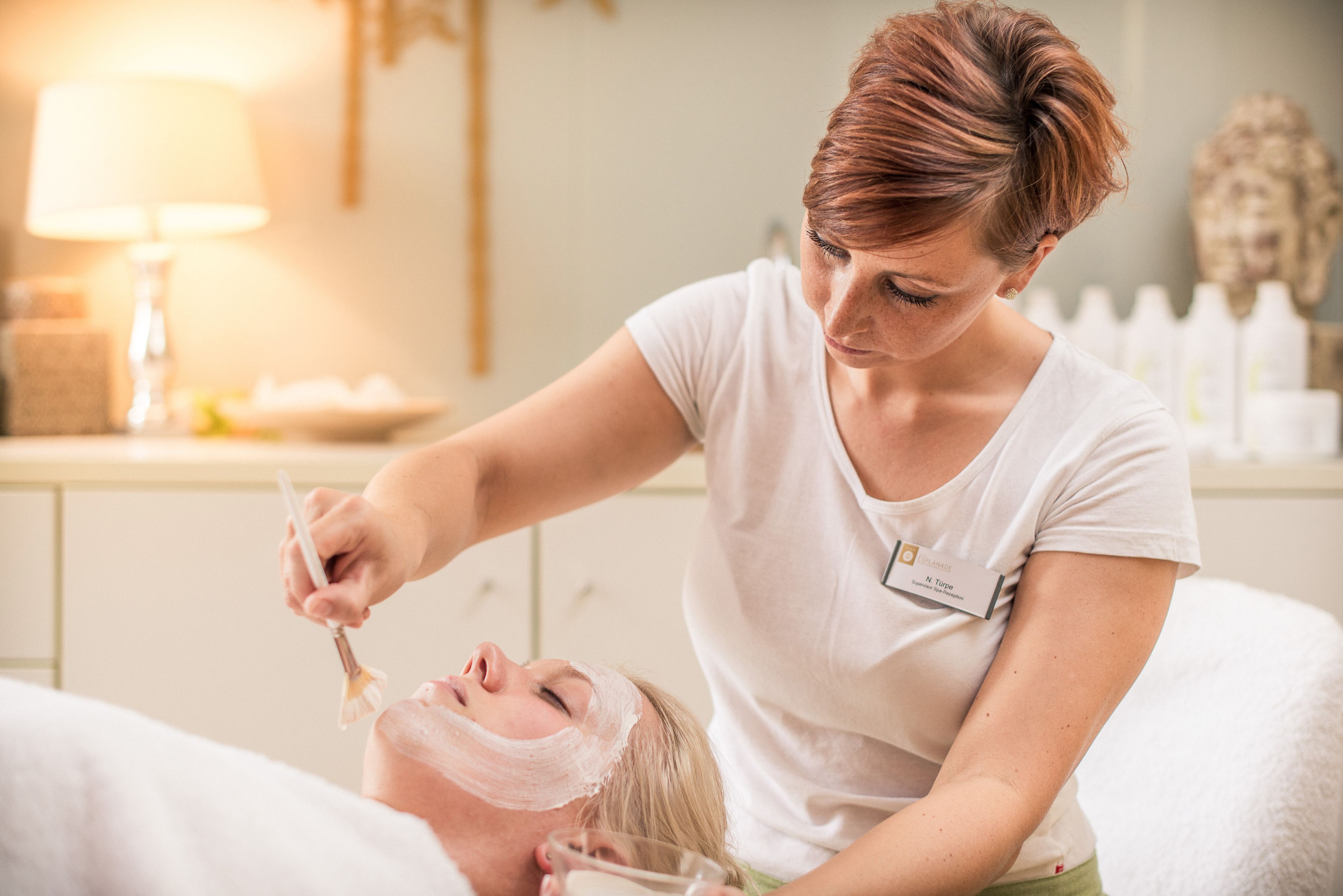 Una persona recibiendo un tratamiento facial en una habitación con una cama blanca, una lámpara y un estante con botellas al fondo.