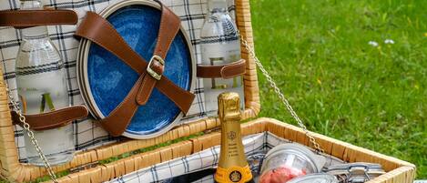 A wicker picnic basket with a plaid cover, containing a bottle, a glass, a bowl, and a plate.