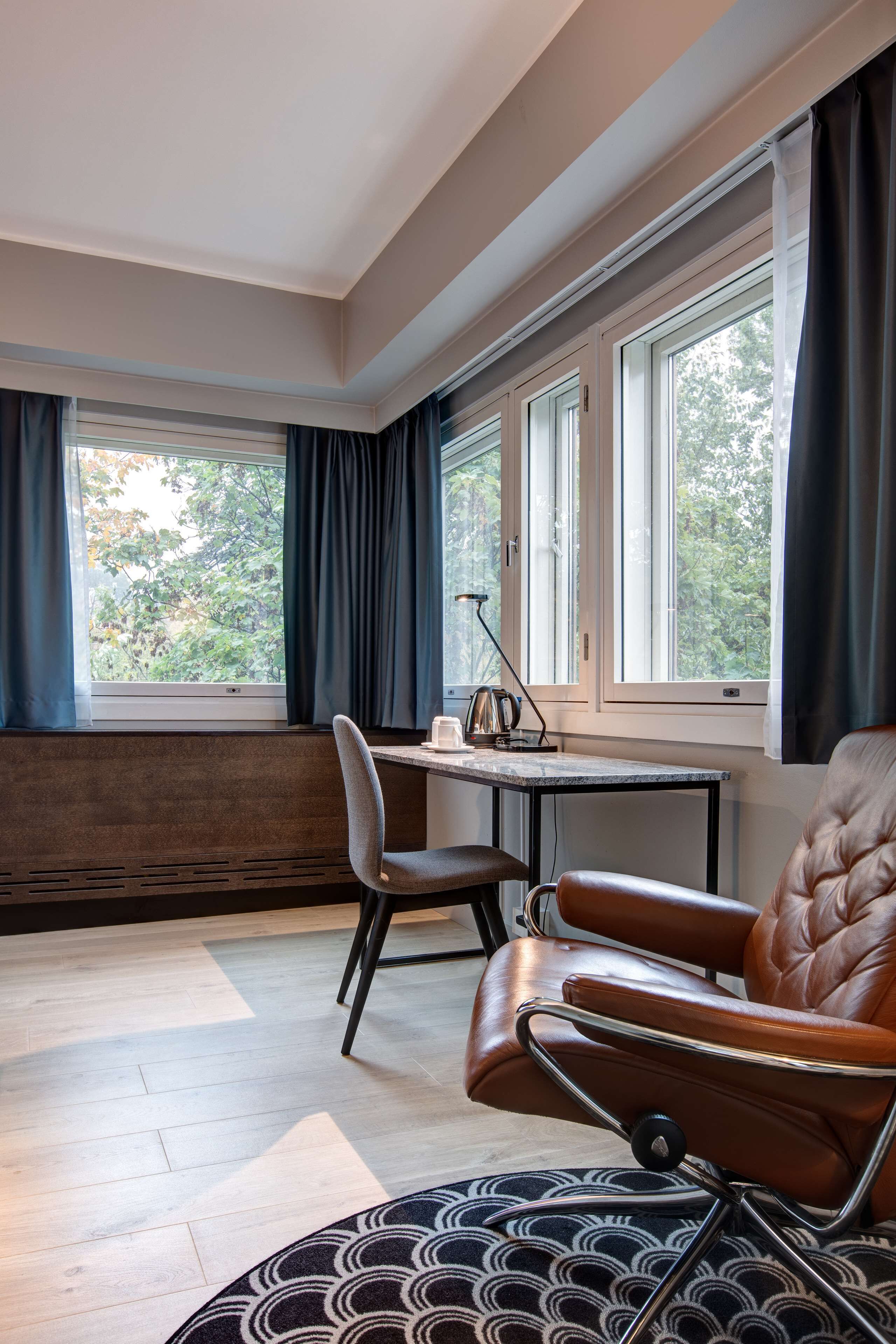 Una habitación moderna con un ventanal grande, una mesa con lámpara, un sillón de cuero y un tapete estampado.