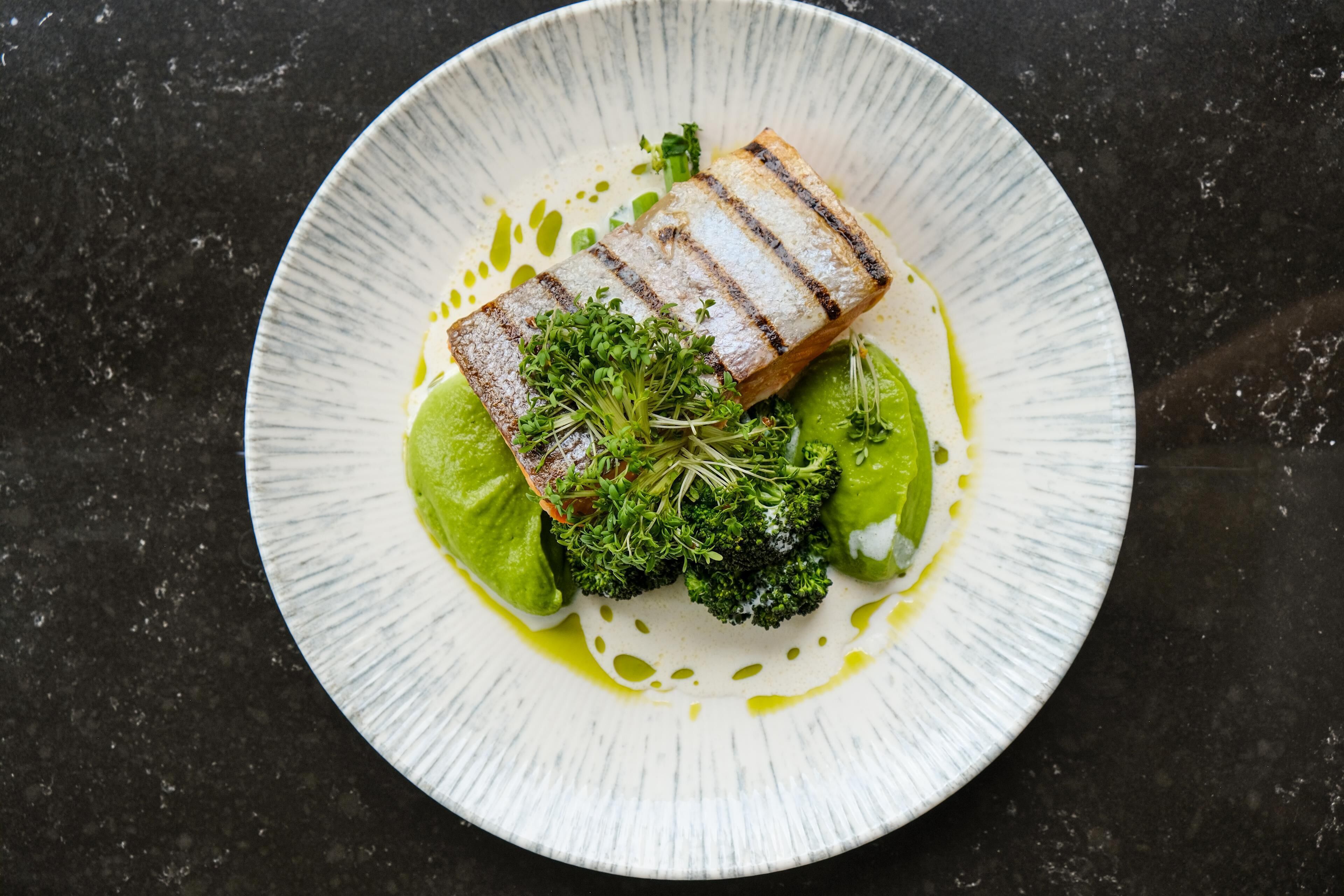 Un plato con pescado a la parrilla, brócoli y salsa verde sobre un plato con textura.