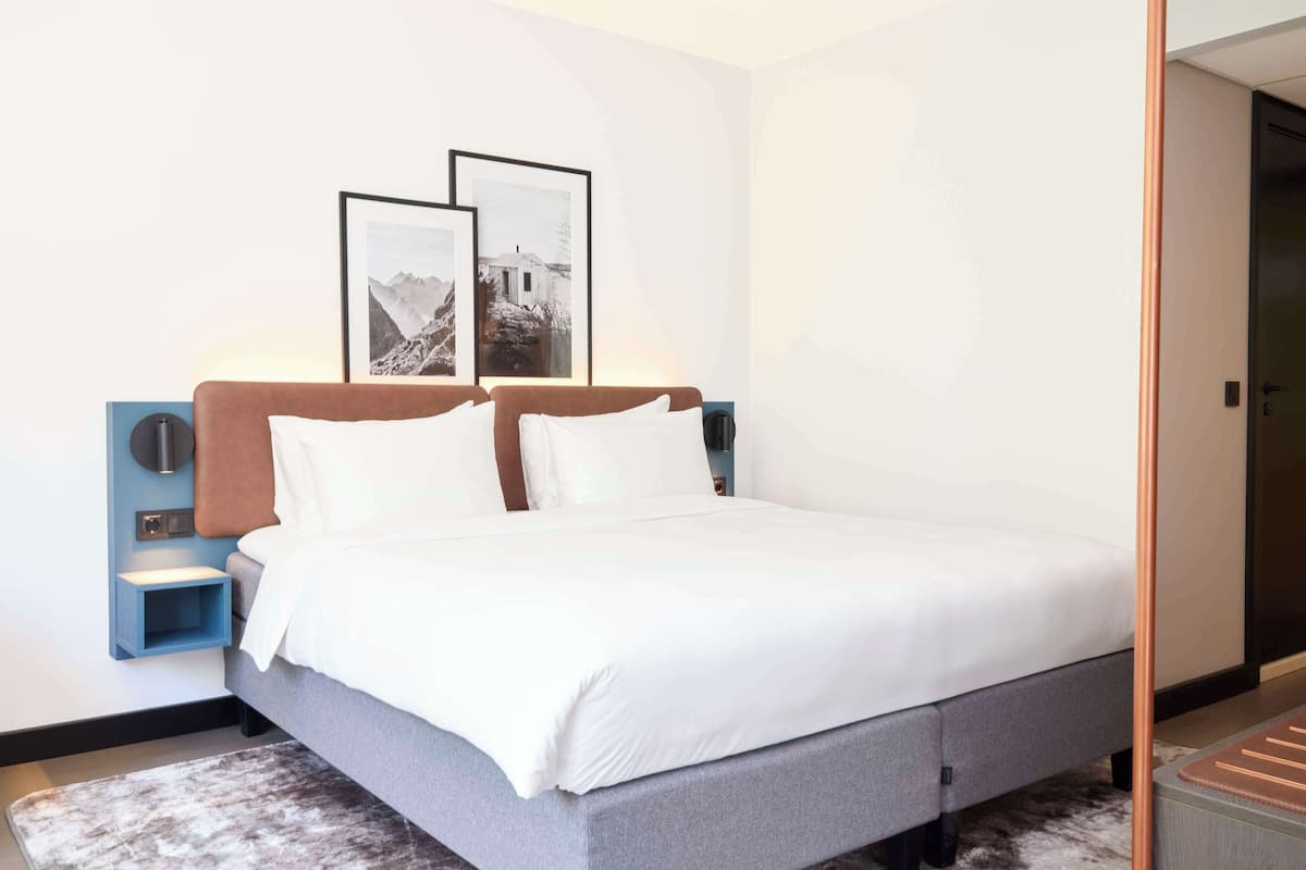 A modern hotel room with a large bed, a nightstand, a wall-mounted lamp, and two framed pictures on the wall.