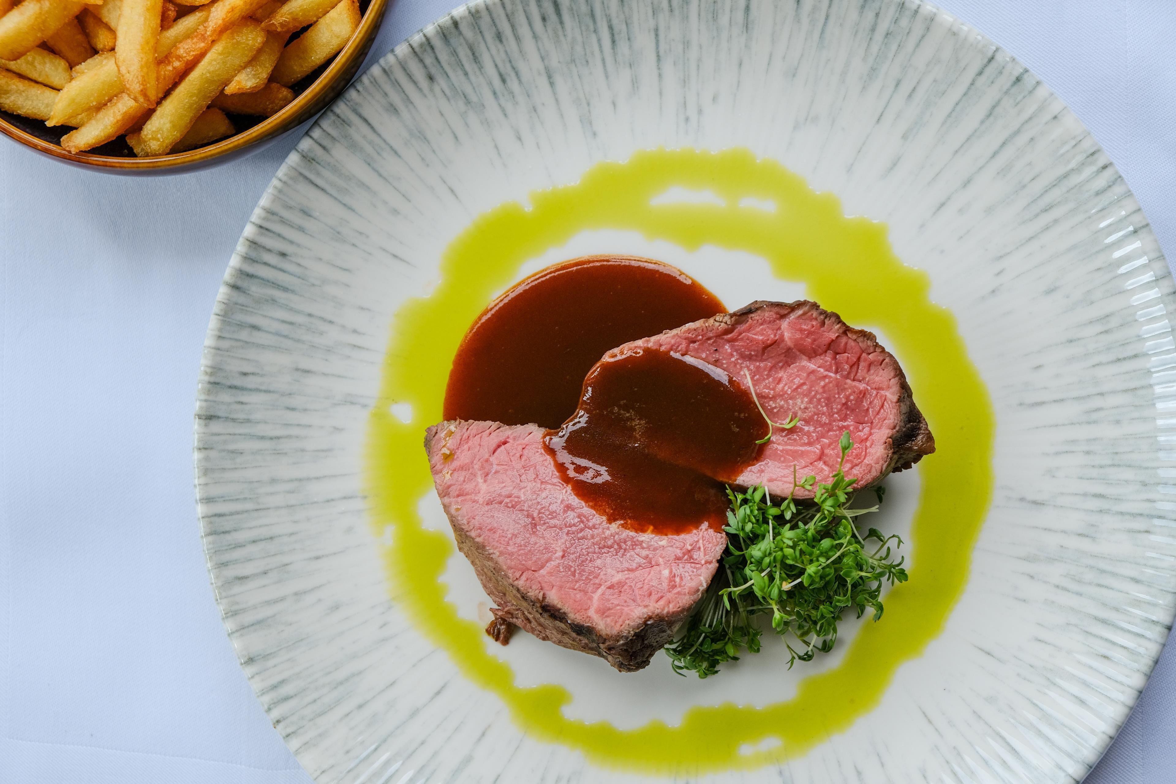 A plate of steak with sauce and greens, and a bowl of fries.