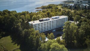 An aerial view of a modern building surrounded by dense greenery, overlooking a large body of water with a distant shoreline. - Radisson Blu Park Hotel, Oslo (Bærum)