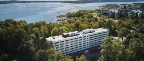 An aerial view of a modern building surrounded by dense greenery, overlooking a large body of water with a distant shoreline.