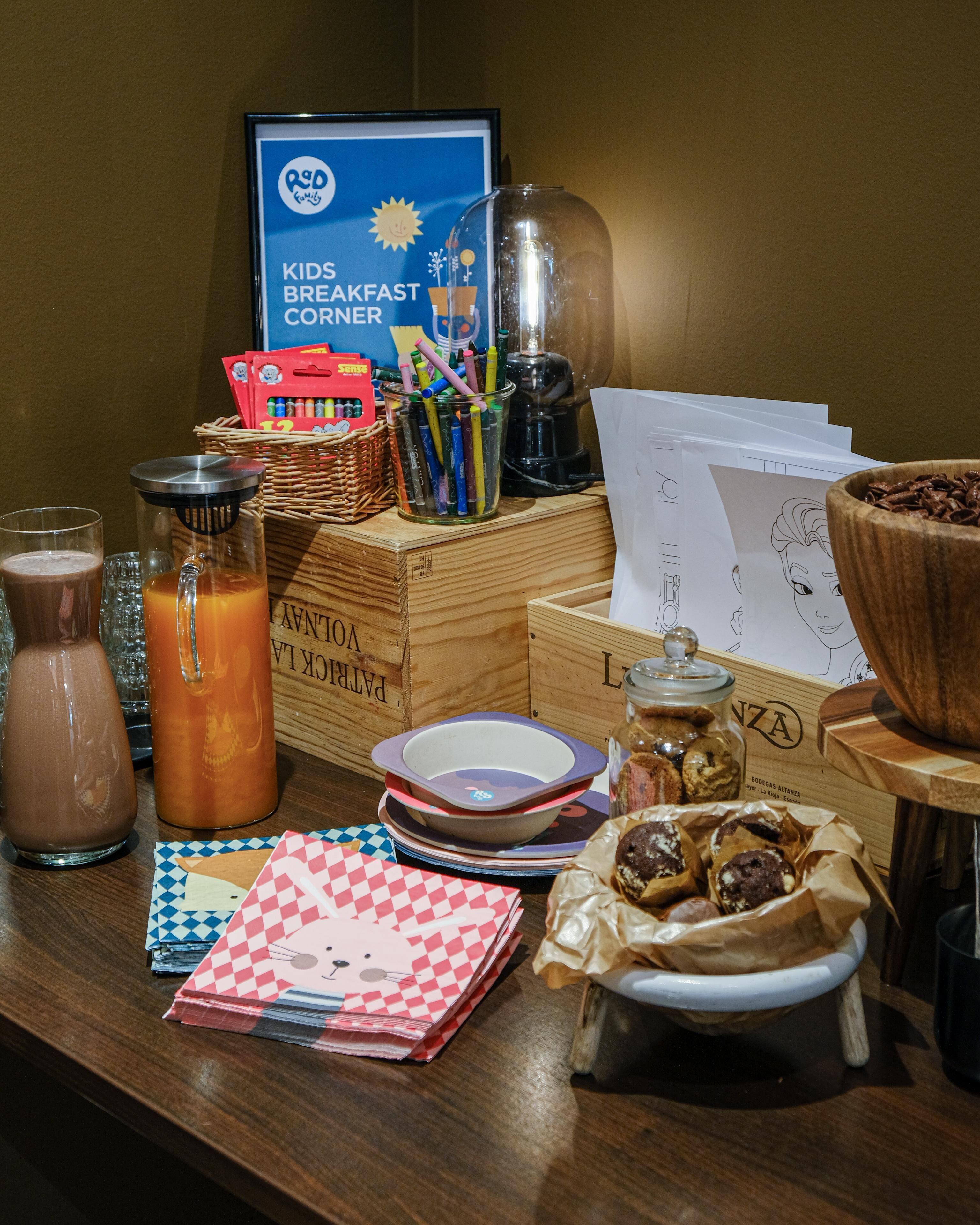A breakfast table with various items including a coffee maker, a basket of crayons, and a sign for a kids breakfast corner.