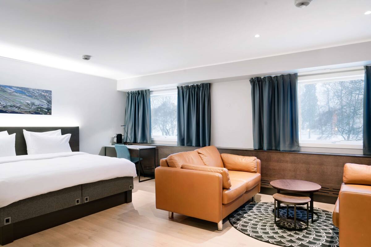 A modern hotel room with a bed, a sofa, a small table, and a view of the outdoors through large windows.