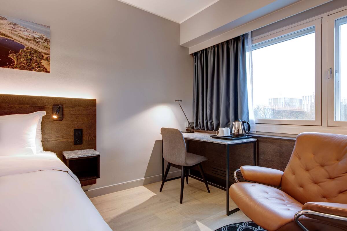 A hotel room with a bed, a desk with a chair, a window with curtains, and a painting on the wall.