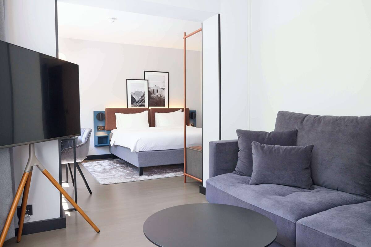 A modern hotel room with a large bed, a grey sofa, a flat-screen TV, and a small side table.