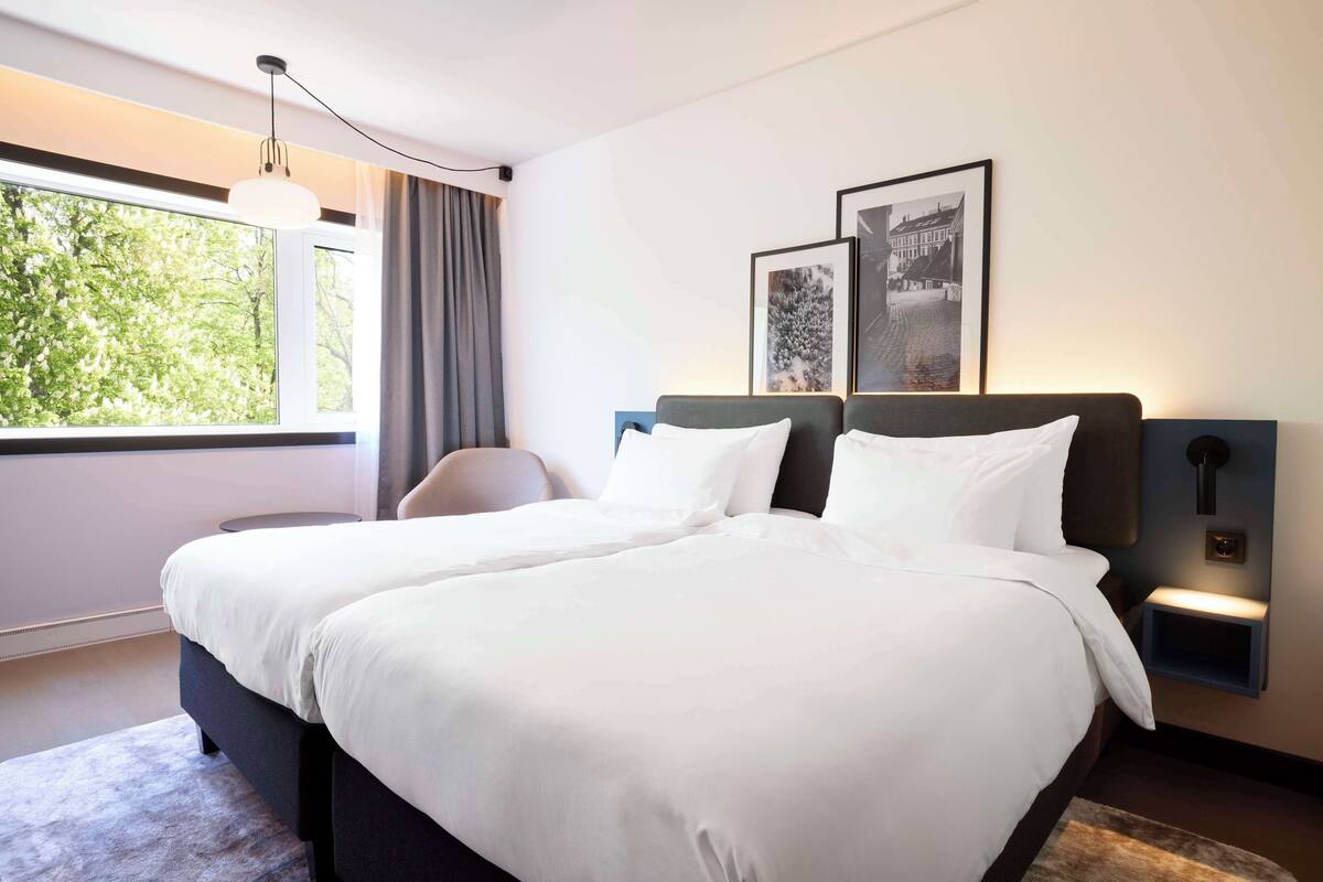 A modern hotel room with a large bed, a desk, and a view of greenery through the window.